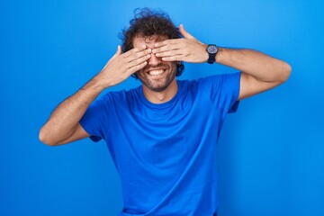 Hispanic young man standing over blue background covering eyes with hands smiling cheerful and funny. blind concept.