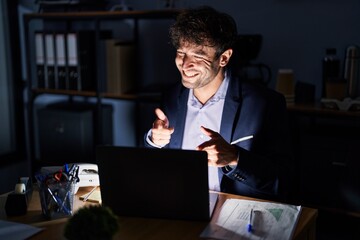 Hispanic young man working at the office at night pointing fingers to camera with happy and funny face. good energy and vibes.