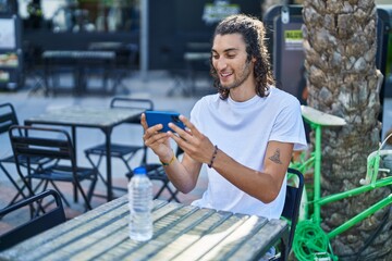 Young hispanic man watching video on smartphone sitting on table at coffee shop terrace