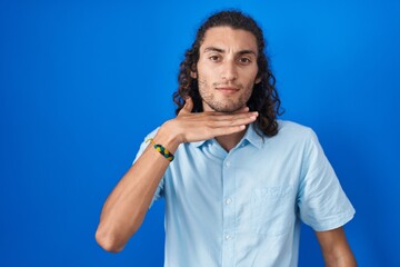 Young hispanic man standing over blue background cutting throat with hand as knife, threaten aggression with furious violence