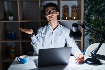 Hispanic man working at the office at night smiling cheerful presenting and pointing with palm of hand looking at the camera.