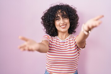 Young middle east woman standing over pink background smiling cheerful offering hands giving assistance and acceptance.