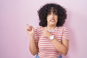 Young middle east woman standing over pink background pointing aside worried and nervous with both hands, concerned and surprised expression