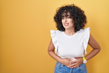Young middle east woman standing over yellow background with hand on stomach because indigestion, painful illness feeling unwell. ache concept.