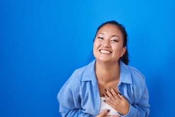 Asian young woman standing over blue background smiling and laughing hard out loud because funny crazy joke with hands on body.