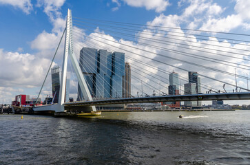 Cityscape of the Dutch city Rotterdam with high rise buildings in the financial district and port area with the Erasmus bridge seen from the water against a blue sky with fluffy clouds.