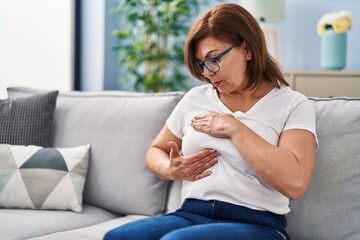 Middle age woman sitting on sofa examining breast with hands at home