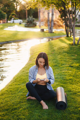 Adult brunette caucasian woman using cellphone while sitting on green grass near yoga mat in park. Mobile phone communication. Sitting woman outdoors. Happy