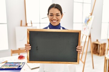 Young latin woman smiling confident holding blackboard at art studio