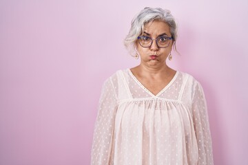 Middle age woman with grey hair standing over pink background puffing cheeks with funny face. mouth...