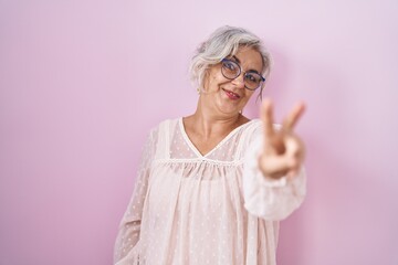 Middle age woman with grey hair standing over pink background smiling looking to the camera showing fingers doing victory sign. number two.