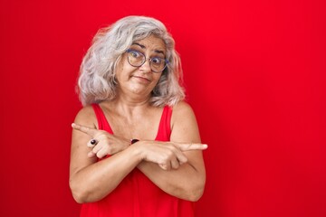 Middle age woman with grey hair standing over red background pointing to both sides with fingers, different direction disagree