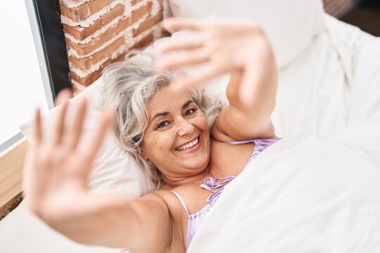Middle age grey-haired woman lying on bed make photo gesture with hands at bedroom