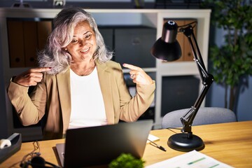 Middle age woman with grey hair working using computer laptop late at night looking confident with smile on face, pointing oneself with fingers proud and happy.