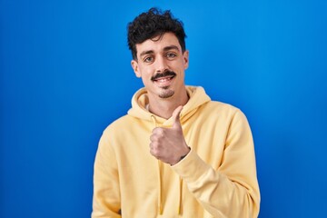 Hispanic man standing over blue background doing happy thumbs up gesture with hand. approving expression looking at the camera showing success.