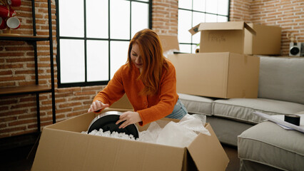 Young redhead woman smiling confident unpacking clock of cardboard box at new home