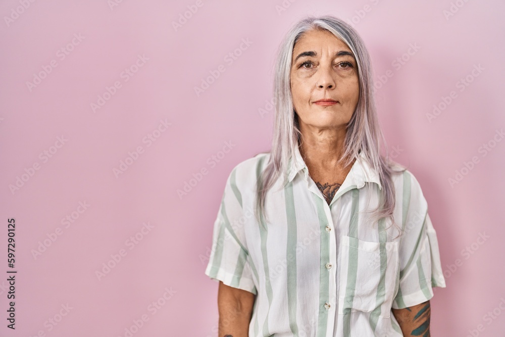 Poster Middle age woman with grey hair standing over pink background with serious expression on face. simple and natural looking at the camera.