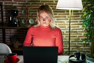 Blonde woman using laptop at night at home with hand on stomach because nausea, painful disease feeling unwell. ache concept.