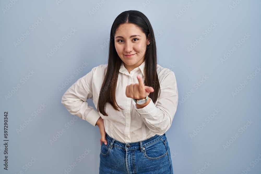 Wall mural Young latin woman standing over blue background beckoning come here gesture with hand inviting welcoming happy and smiling