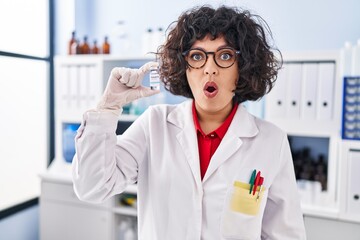 Hispanic doctor woman with curly hair holding vaccine scared and amazed with open mouth for surprise, disbelief face