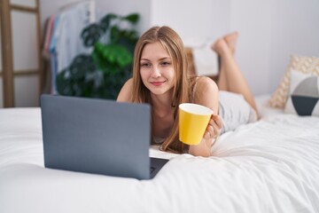 Obraz na płótnie Canvas Young caucasian woman using laptop drinking coffee lying on bed at bedroom