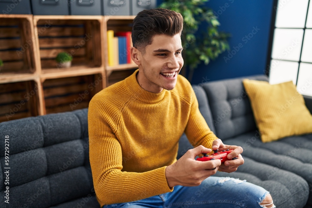 Wall mural young hispanic man playing video game sitting on sofa at home