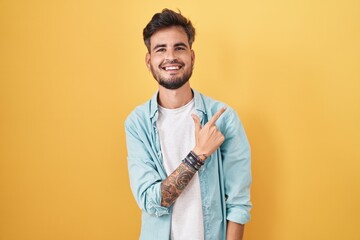Young hispanic man with tattoos standing over yellow background cheerful with a smile on face...