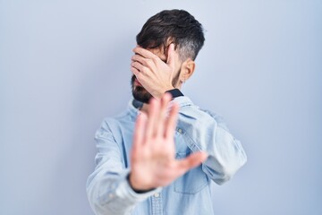 Young hispanic man with beard standing over blue background covering eyes with hands and doing stop gesture with sad and fear expression. embarrassed and negative concept.