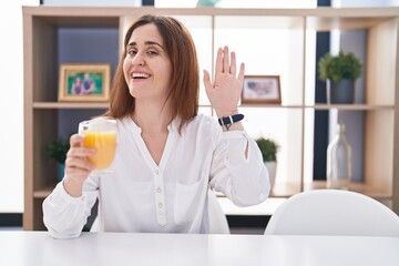 Brunette woman drinking glass of orange juice waiving saying hello happy and smiling, friendly welcome gesture