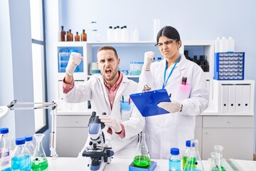 Two young people working at scientist laboratory annoyed and frustrated shouting with anger, yelling crazy with anger and hand raised