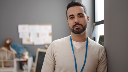 Young hispanic man preschool teacher standing with relaxed expression at kindergarten