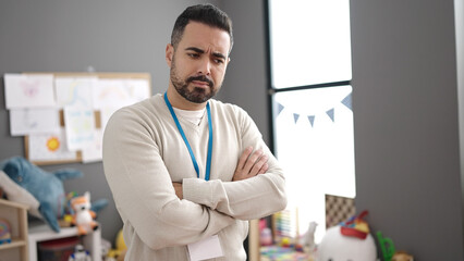 Young hispanic man teacher standing with arms crossed gesture at kindergarten