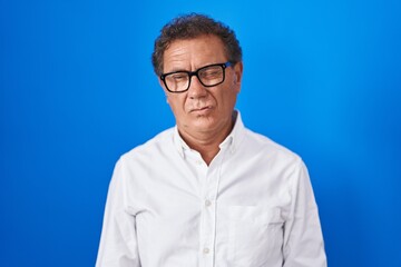 Middle age hispanic man standing over blue background winking looking at the camera with sexy expression, cheerful and happy face.