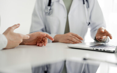 Doctor and patient sitting at the desk in clinic office. The focus is on female physician's hands pointing to laptop computer monitor, close up. Perfect medical service and medicine concept.