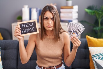 Young hispanic woman holding blackboard with new home text and dollars skeptic and nervous,...