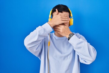 Non binary person listening to music using headphones covering eyes and mouth with hands, surprised and shocked. hiding emotion