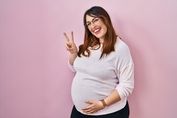Pregnant woman standing over pink background showing and pointing up with fingers number two while smiling confident and happy.