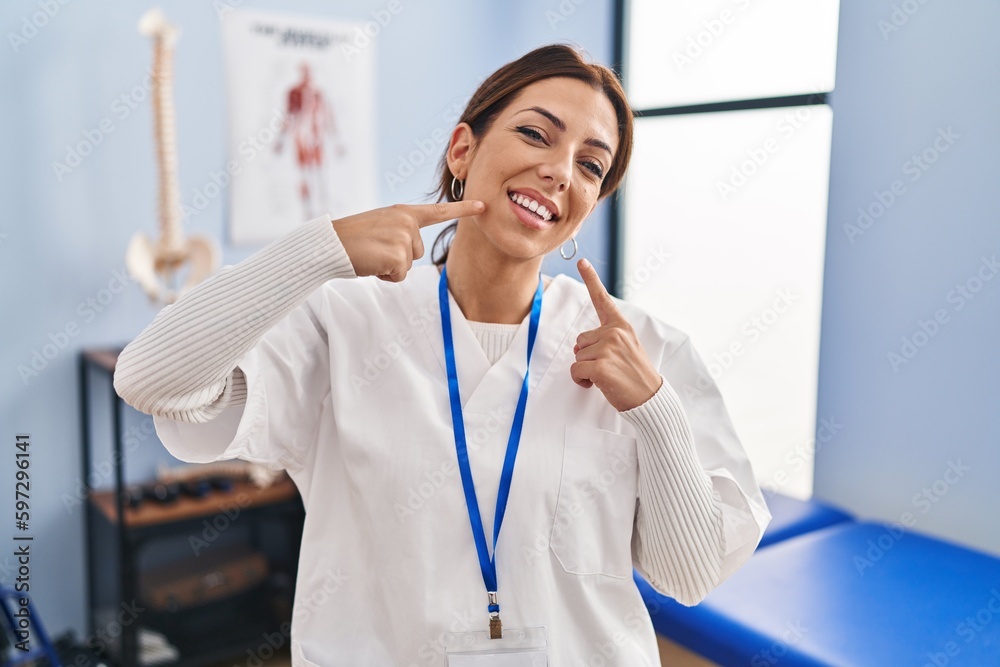 Sticker Young brunette woman working at pain recovery clinic smiling cheerful showing and pointing with fingers teeth and mouth. dental health concept.