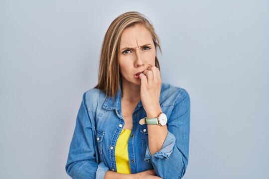 Young blonde woman standing over blue background looking stressed and nervous with hands on mouth biting nails. anxiety problem.