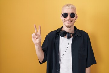 Young caucasian man wearing sunglasses standing over yellow background showing and pointing up with fingers number two while smiling confident and happy.