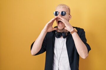 Young caucasian man wearing sunglasses standing over yellow background shouting angry out loud with hands over mouth