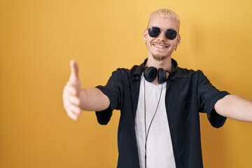 Young caucasian man wearing sunglasses standing over yellow background looking at the camera smiling with open arms for hug. cheerful expression embracing happiness.