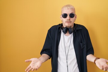 Young caucasian man wearing sunglasses standing over yellow background clueless and confused with open arms, no idea concept.