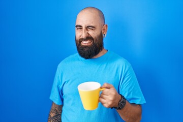 Young hispanic man with beard and tattoos drinking a cup of coffee winking looking at the camera with sexy expression, cheerful and happy face.