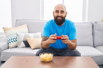 Young hispanic man with beard and tattoos playing video game sitting on the sofa smiling with a happy and cool smile on face. showing teeth.