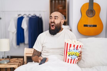 Young hispanic man with beard and tattoos eating popcorn in the bed angry and mad screaming...