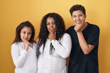Family of mother, daughter and son standing over yellow background with hand on chin thinking about...