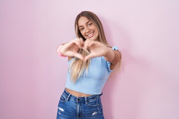 Young blonde woman standing over pink background smiling in love doing heart symbol shape with hands. romantic concept.