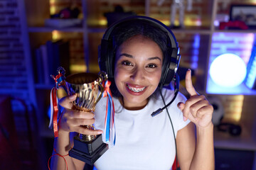 Young arab woman playing video games holding trophy smiling with an idea or question pointing...
