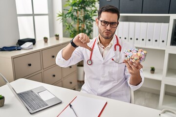Young hispanic doctor man with beard holding candy with angry face, negative sign showing dislike with thumbs down, rejection concept